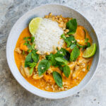 Grey stone table with a grey ceramic bowl filled with Chicken Pumpkin Thai Red Curry and topped with mint, basil, chopped peanuts and lime wedges