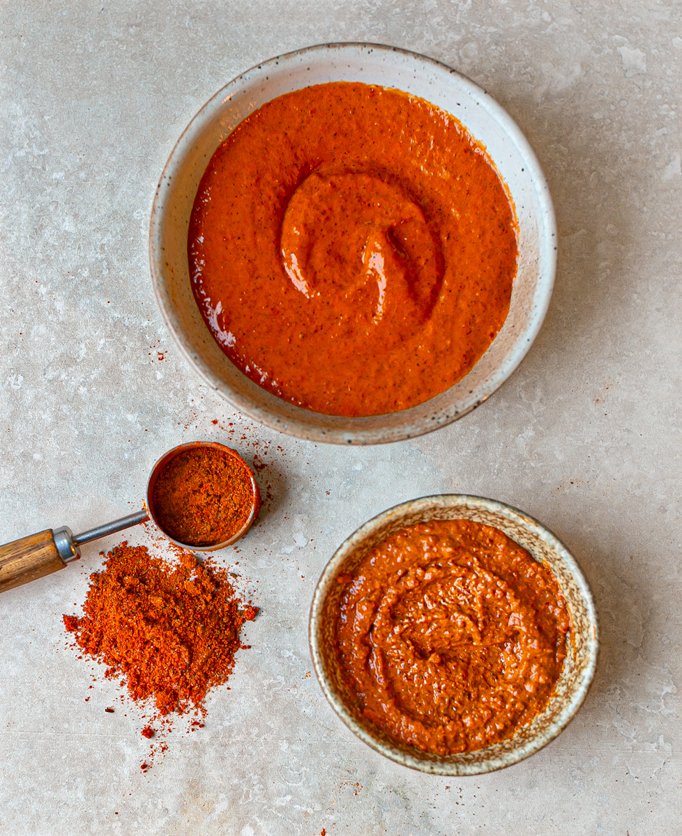 Harissa powder, paste, and sauce displayed on a light stone surface. The image shows vibrant red harissa powder in a measuring spoon with a small pile beside it, along with two bowls containing smooth harissa paste and a slightly chunky harissa sauce. Perfect for adding bold, spicy North African flavour to dishes