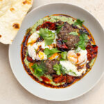 beige ceramic bowl filled with fusion turkish eggs of charred green feta sauce and topped with poached eggs, beef kofta, fresh herbs and flat breads