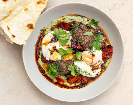 beige ceramic bowl filled with fusion turkish eggs of charred green feta sauce and topped with poached eggs, beef kofta, fresh herbs and flat breads