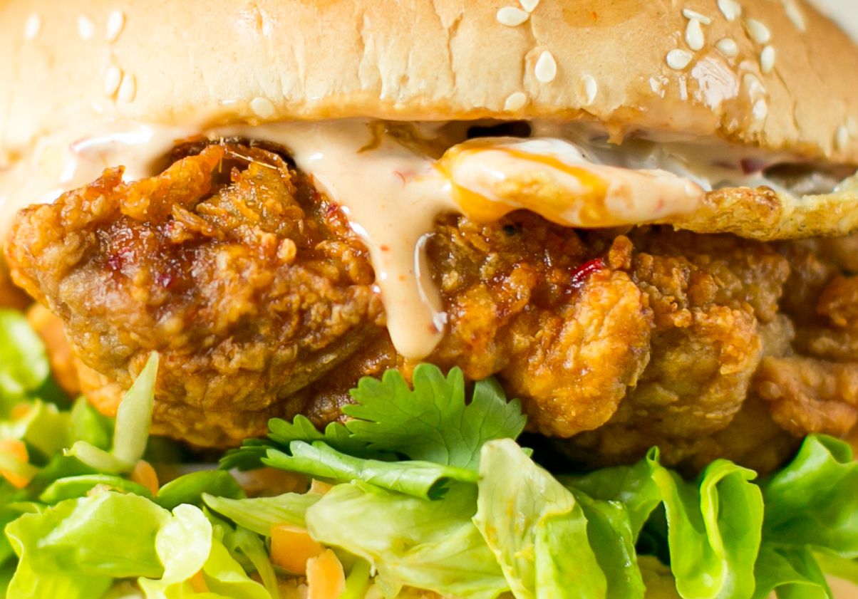 A close up view of a burger with sesame seed bun, crispy fried chicken, with green lettuce, and a reddish spicy mayonnaise sauce dripping down the burger 