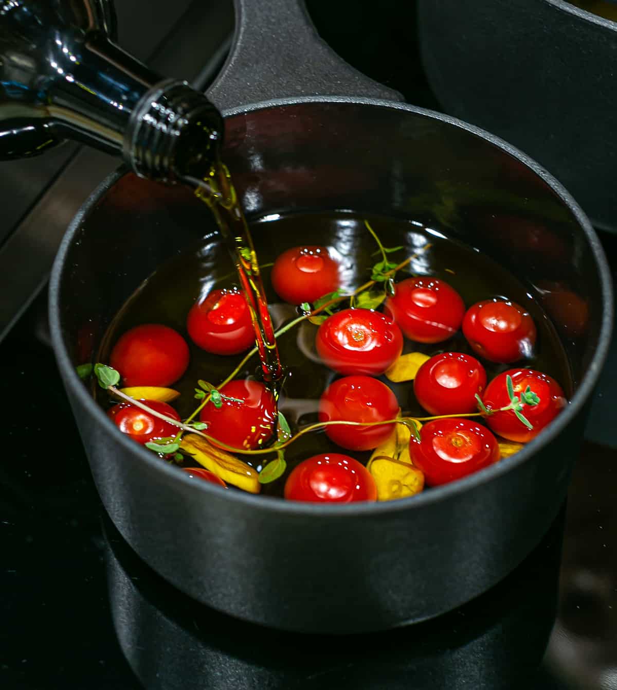 Olive oil being poured into a black stove top saucepan filled cherry tomatoes, garlic cloves and twigs of thyme