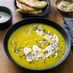 a black soup bowl filled with a bright yellow curried cauliflower soup topped with yoghurt and almonds next to a bowl of garlic nam bread and poppadoms on a copper coloured table
