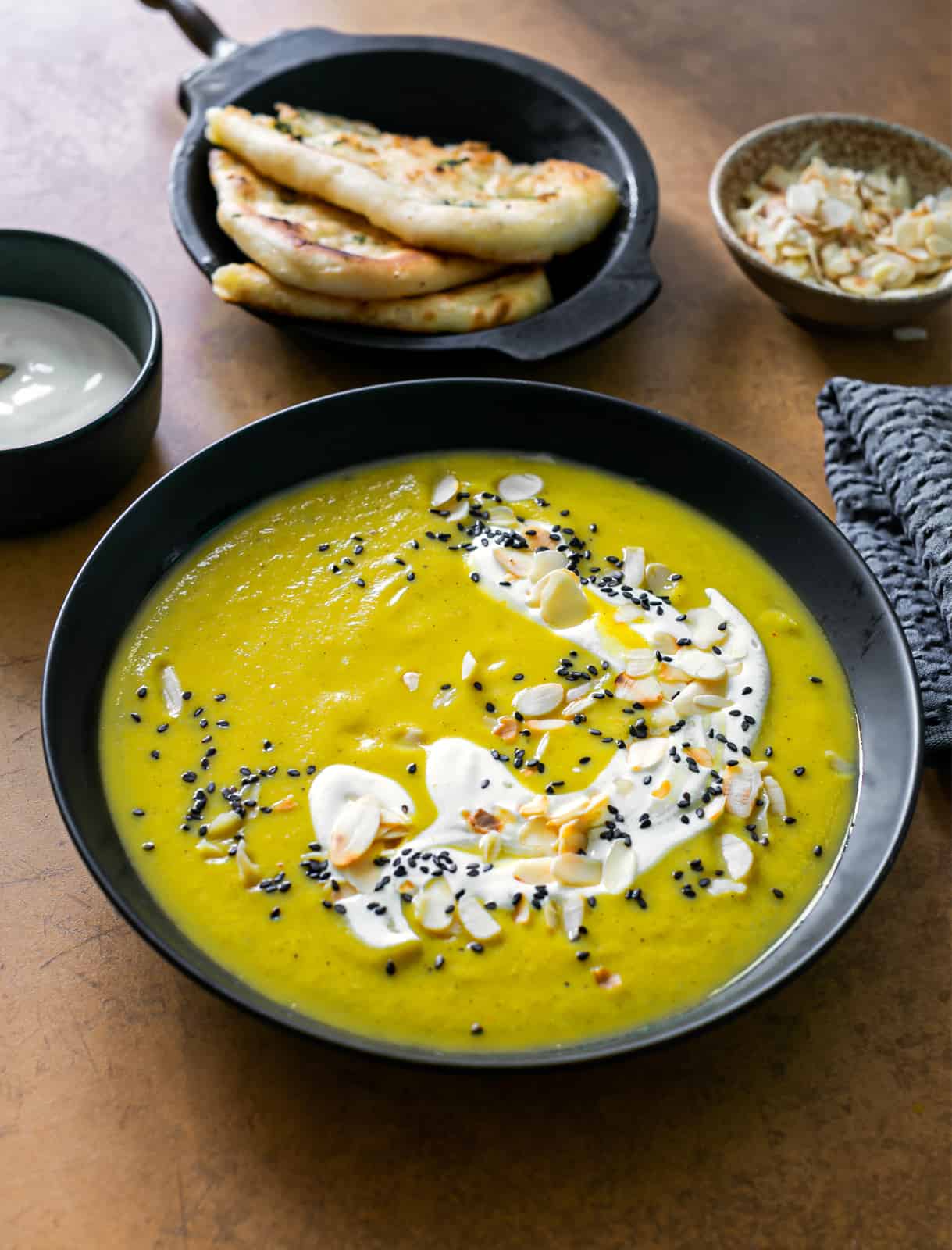 a black soup bowl filled with a bright yellow curried cauliflower soup topped with yoghurt and almonds next to a bowl of garlic nam bread and popadoms on a copper coloured table 