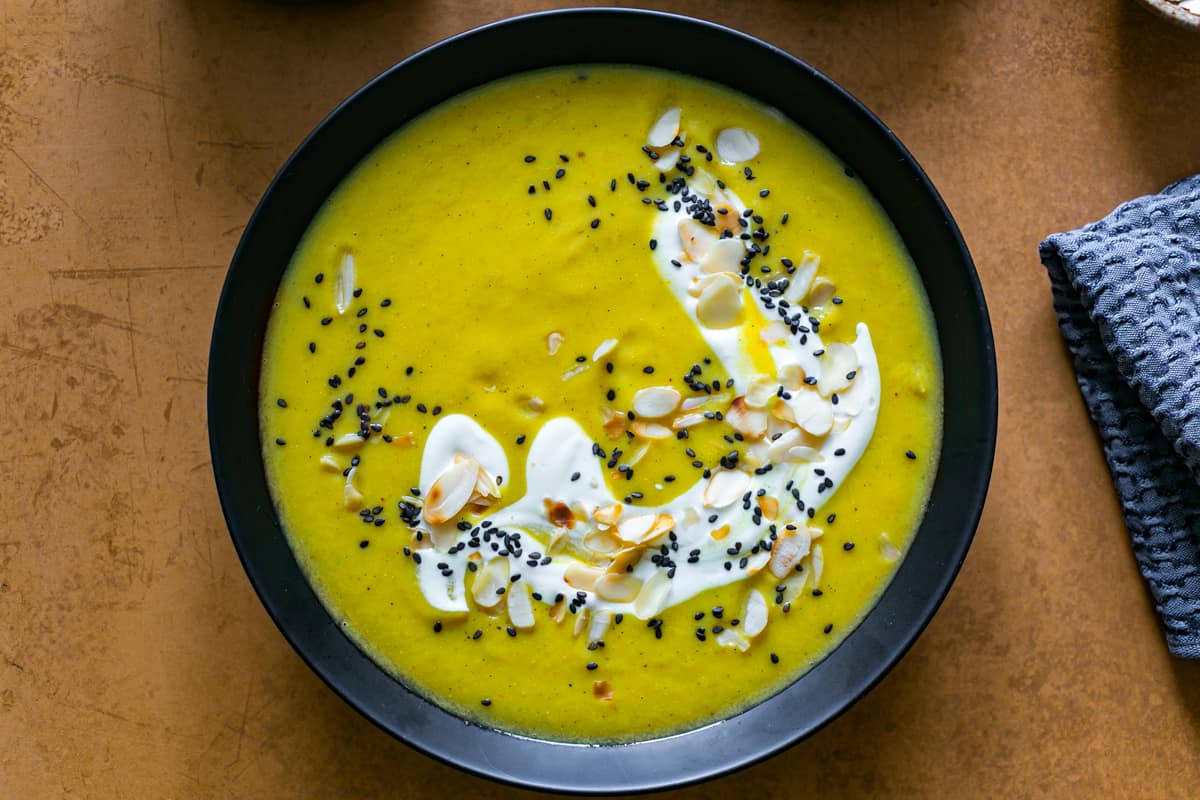 a black soup bowl filled with a bright yellow curried cauliflower soup topped with yoghurt and almonds on a copper coloured table