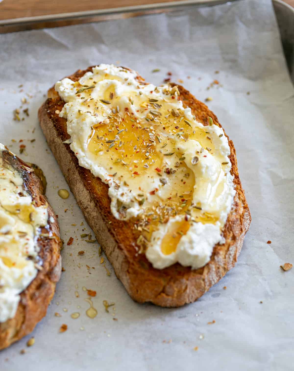 ricotta, honey and spices spread over crusty toasted bread, sitting on white parchment paper and a silver serving tray on top of a brown wooden kitchen top 