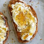 ricotta, honey and spices spread over crusty toasted bread, sitting on white parchment paper and a silver serving tray on top of a brown wooden kitchen top