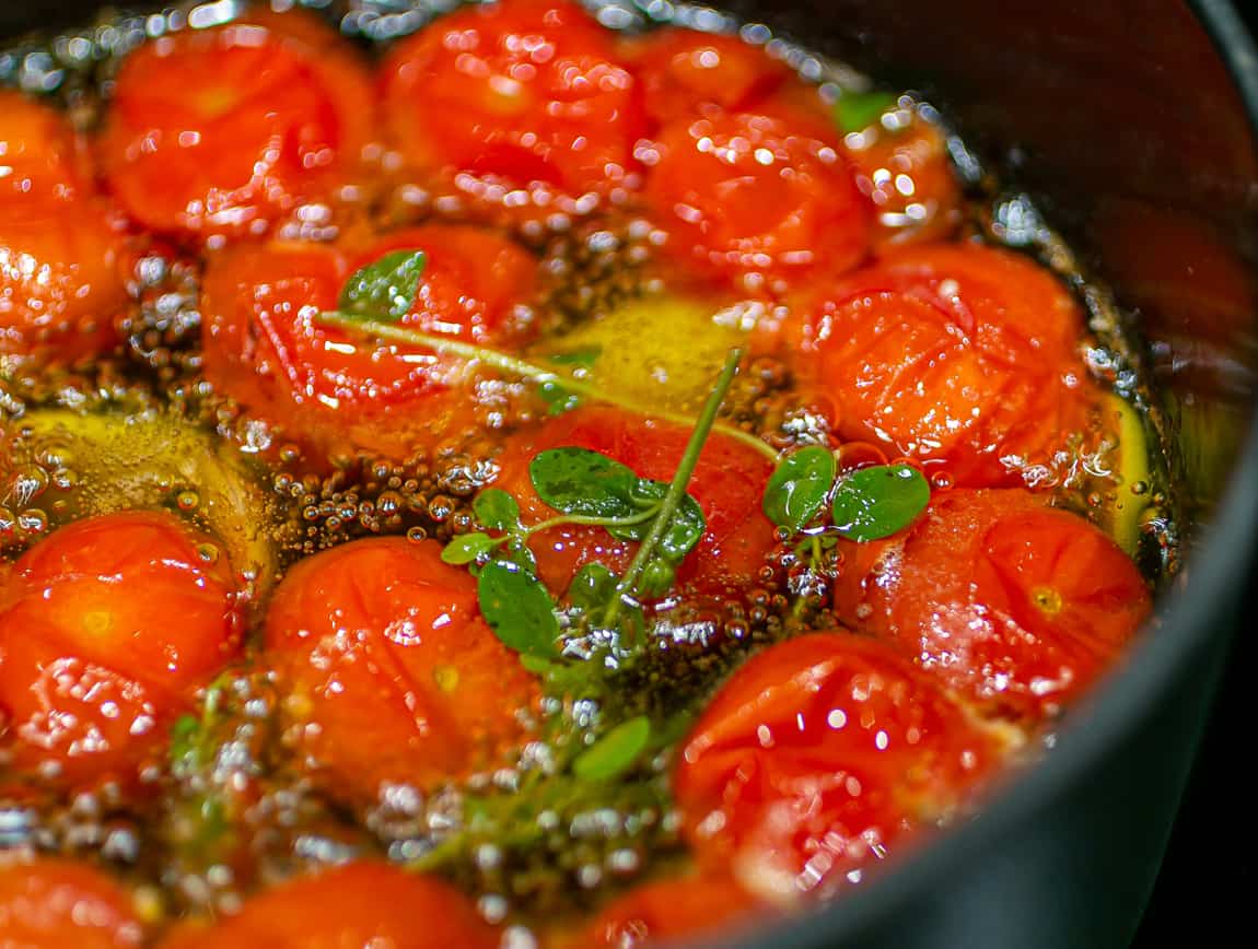 small bright red cherry tomatoes, garlic cloves and a sprig of thyme simmering in a black cast iron pot of olive oil