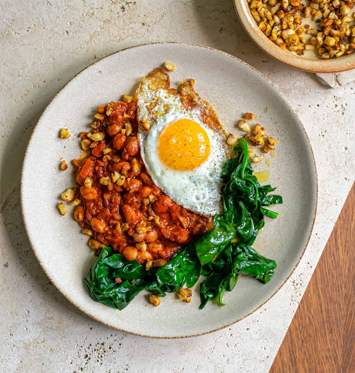 Beige ceramic plate on a beige ceramic table with spicy Italian 'Nduja beans, spinach and a crispy fried egg with bright yellow yolk 