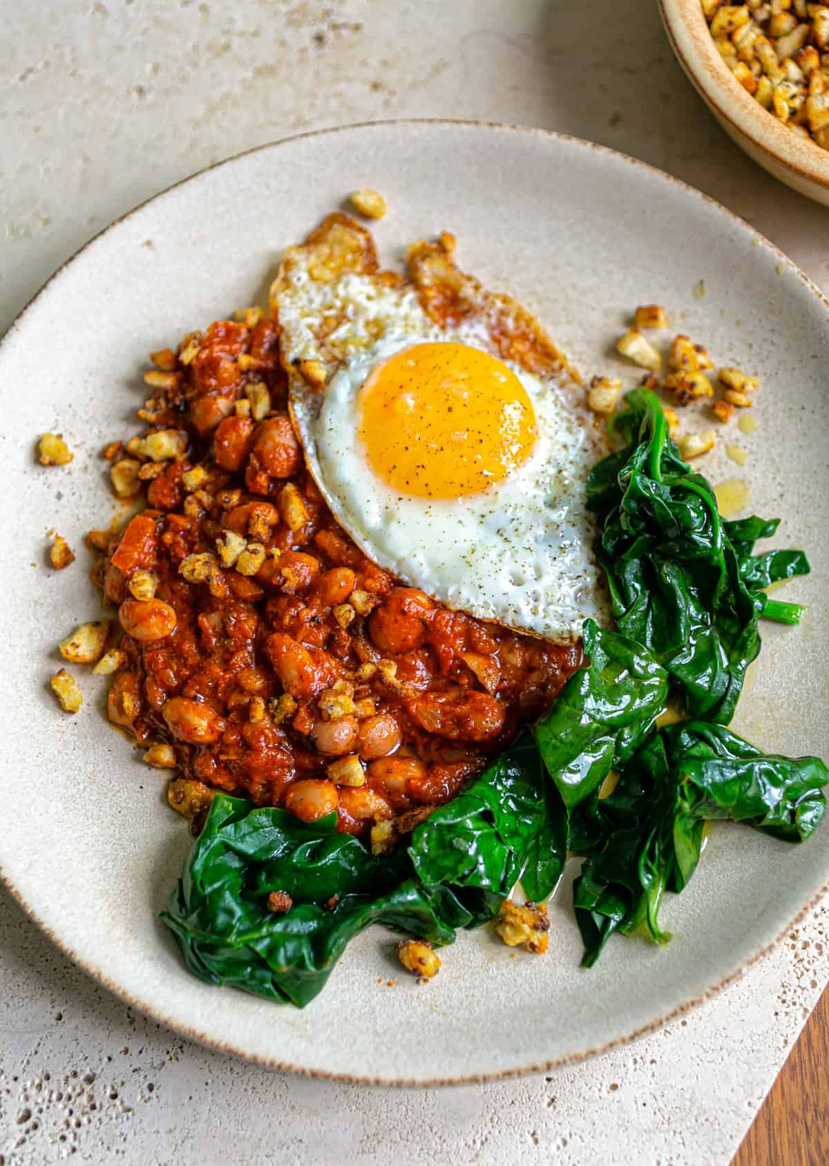 Beige ceramic plate on a beige ceramic table with spicy Italian 'Nduja beans, spinach and a crispy fried egg with bright yellow yolk 
