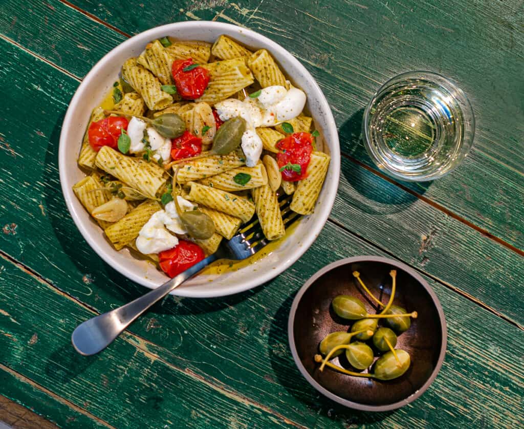 green wooden table with a cream ceramic pasta bowl filled with thick rigatoni pasta, green basil pesto, red confit cherry tomatoes, creamy burrata cheese, caper berries and a glass of white wine 