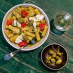 green wooden table with a cream ceramic pasta bowl filled with thick rigatoni pasta, green basil pesto, red confit cherry tomatoes, creamy burrata cheese, caper berries and a glass of white wine