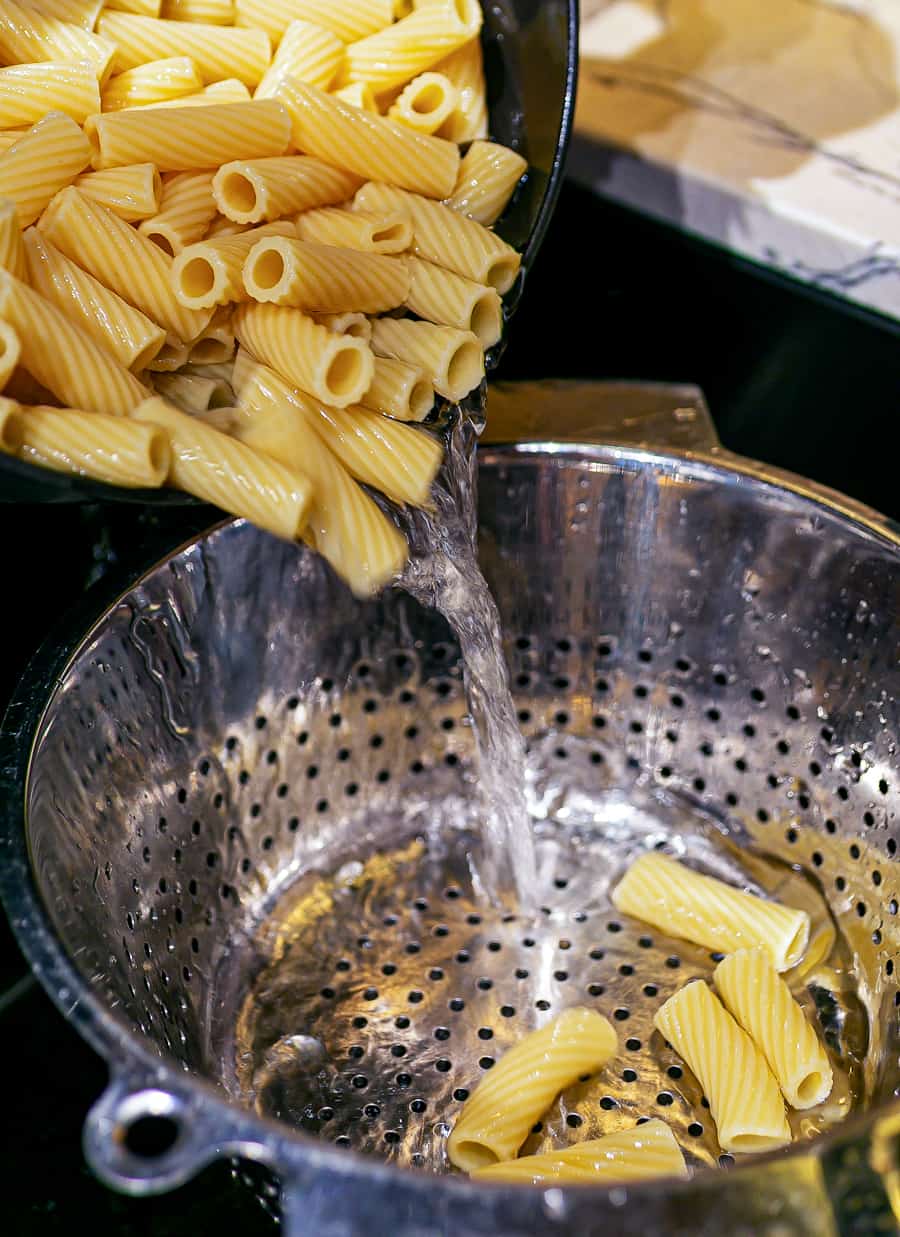 a pot of cooked rigatoni pasta being poured into a large sieve to separate from the cooking water