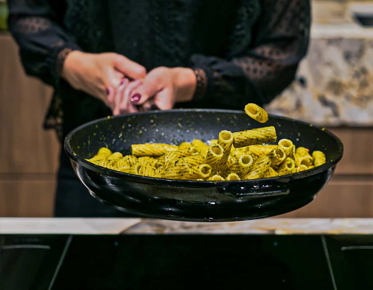A lady wearing a black top stands in front of a kitchen hob holding a large frying pan with rigatoni pasta coated in green pesto being tossed into the air