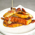 A plate of Maple Cardamom Butter French Toast topped with banana and pecan nuts being drizzled with butter maple sauce