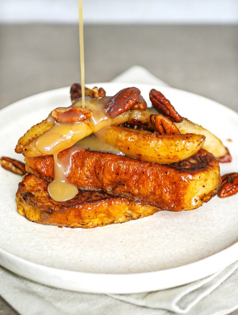 A plate of Maple Cardamom Butter French Toast topped with banana and pecan nuts being drizzled with butter maple sauce