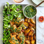 a tray of roasted chicken and potatoes with green salsa verde on a white marble table