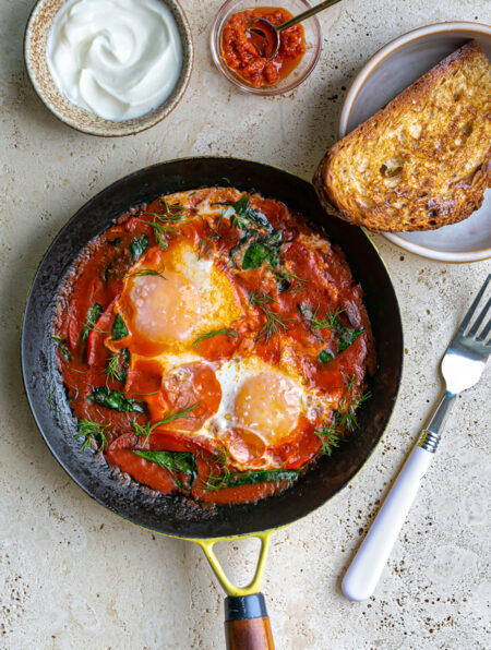 Table with a frying pan filled with Shakshuka and a side of toast, yoghurt and chili sauce