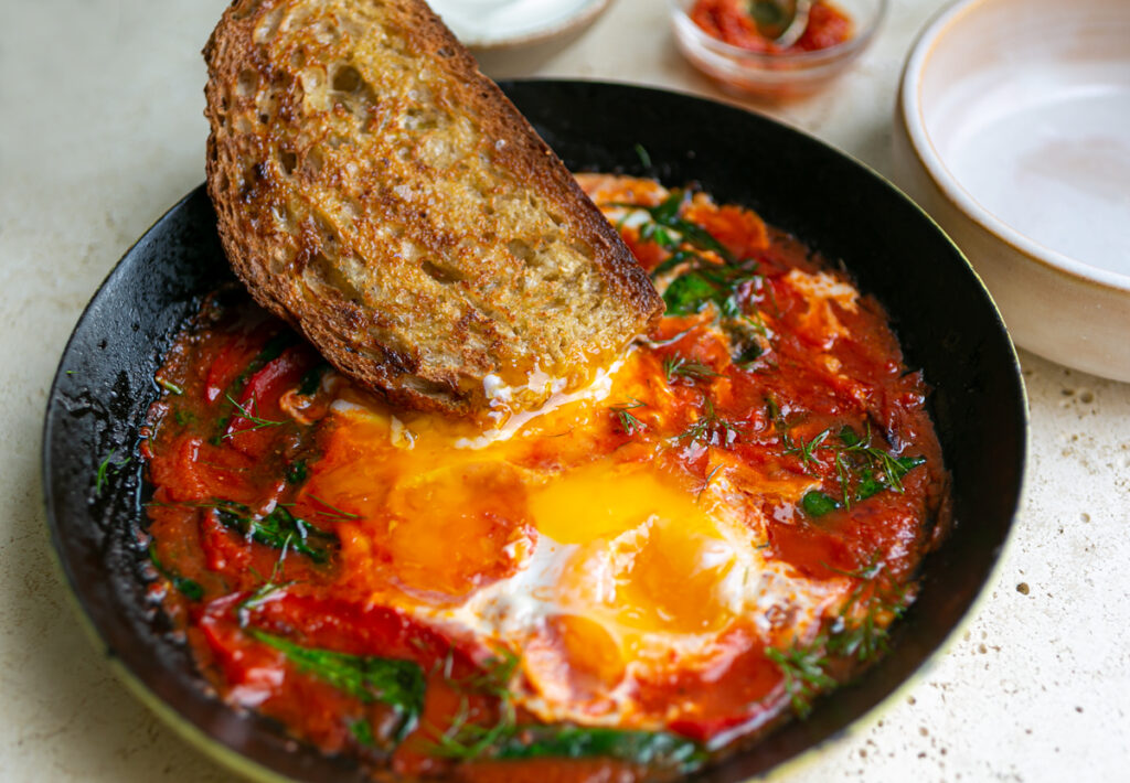 A piece of toast being dipped into runny egg yolks in a pan of shakshuka