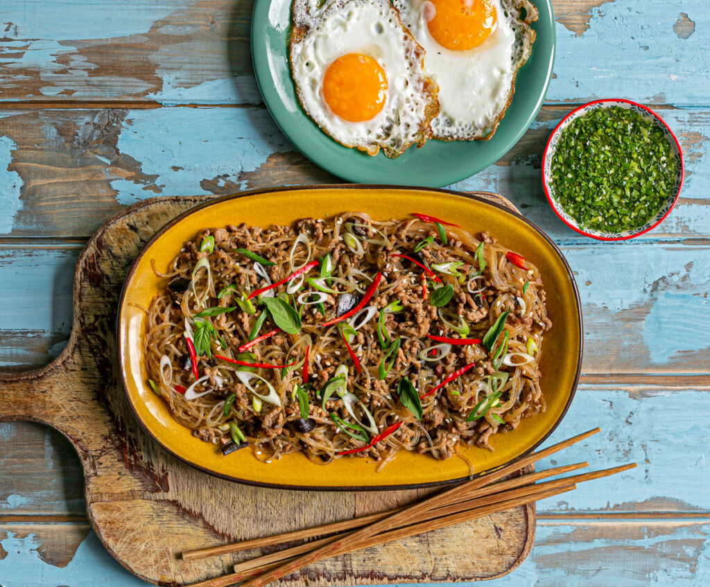 yellow plate on a blue wood table with Asian noodle and meat sauce and fried eggs