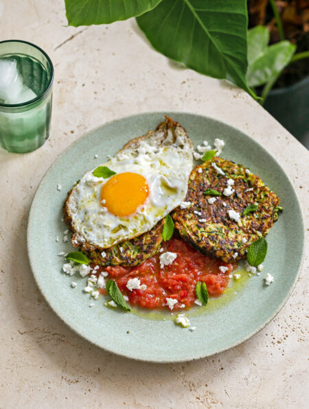 green plate topped with tomato salsa, courgette fritters, a crispy fried egg, garnished with crumbled feta and mint leaves and a glass of iced water