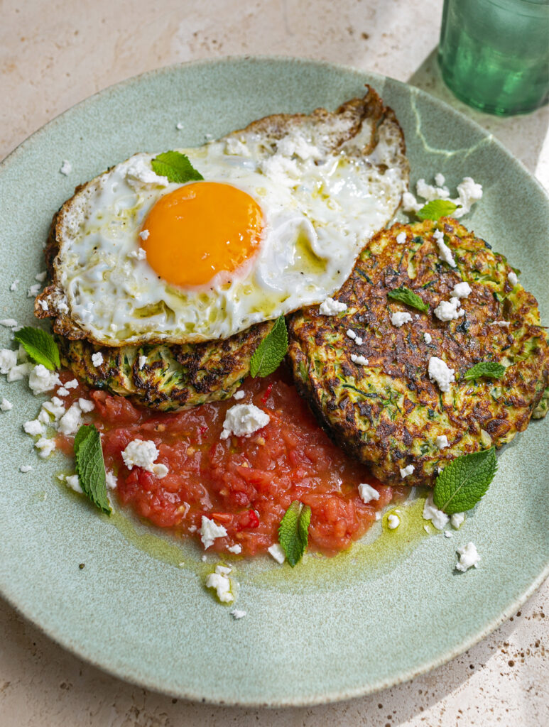 green plate topped with tomato salsa, courgette fritters, a crispy fried egg, garnished with crumbled feta and mint leaves