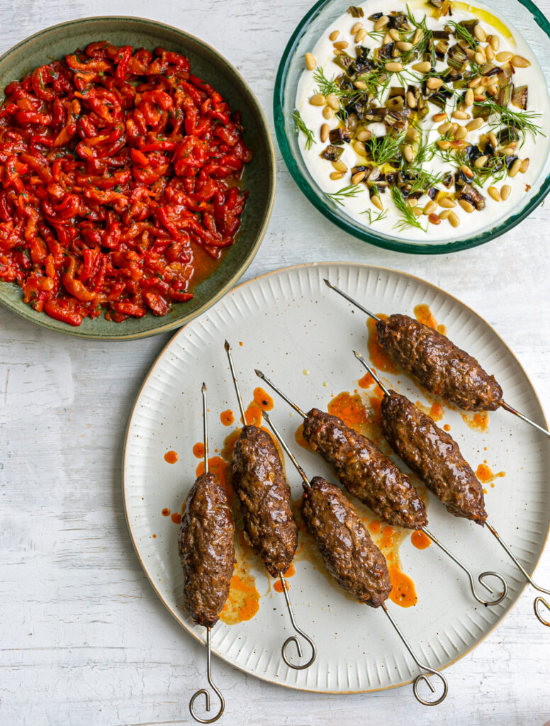 plates on a white wooden table top with beef kebab skewers, roasted red pepper salad and creamy yoghurt feta dip topped with pine nuts and dill fonds