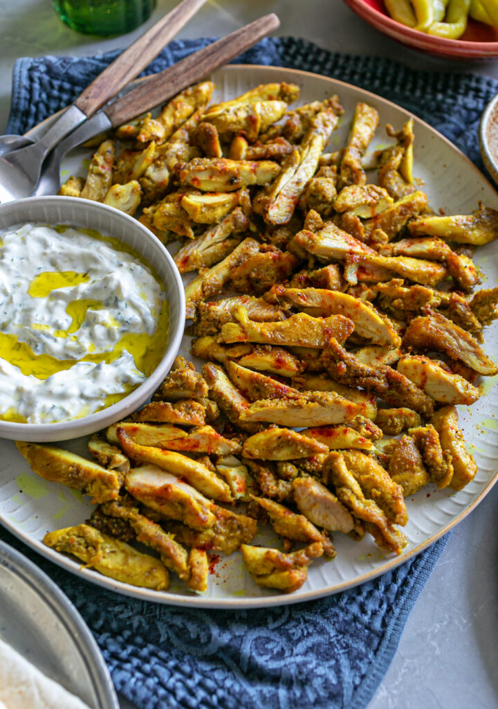 A ceramic beige plate piled with shredded Chicken shawarma pieces an tzatziki