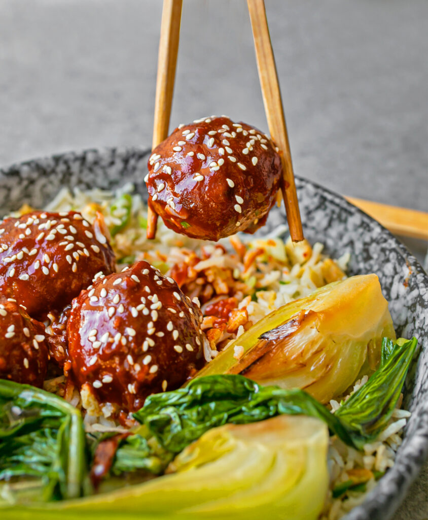 one gochujang meatball held in a chopstick being raised above a blue ceramic bowl of more meatballs, egg fried rice and bok choy 