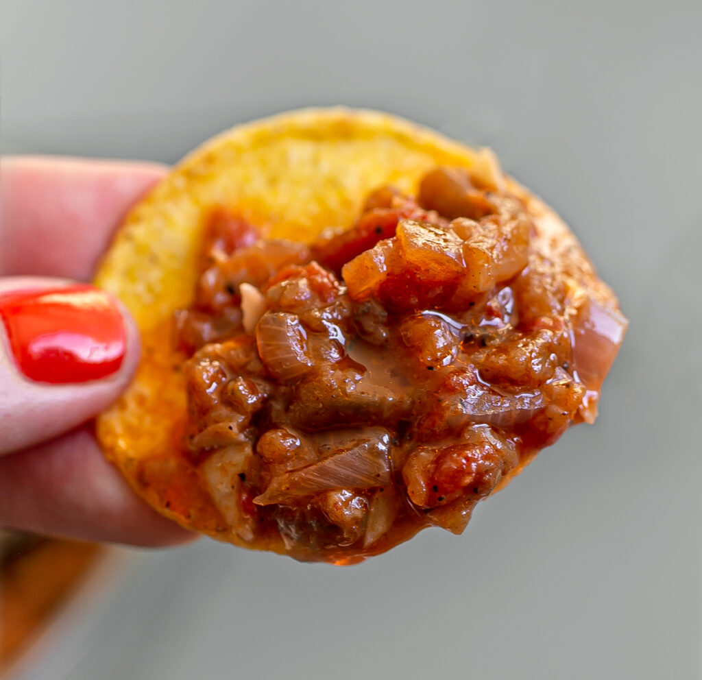 Close up of a hand holding a nacho chip dipped in Aubergine Caviar Bolognese sauce
