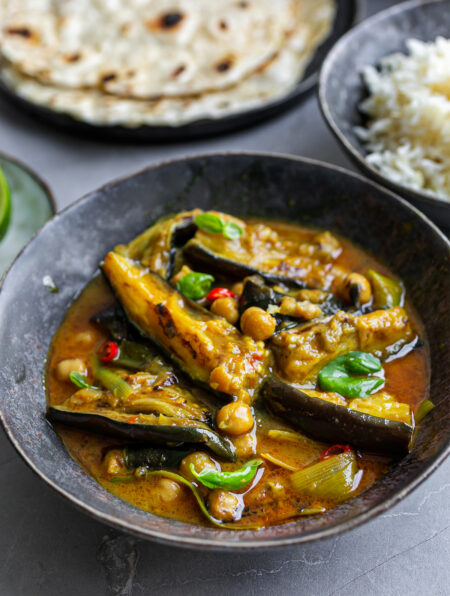 Grey ceramic bowl filled with Fragrant Aubergine Coconut Curry next to rice and flatbreads