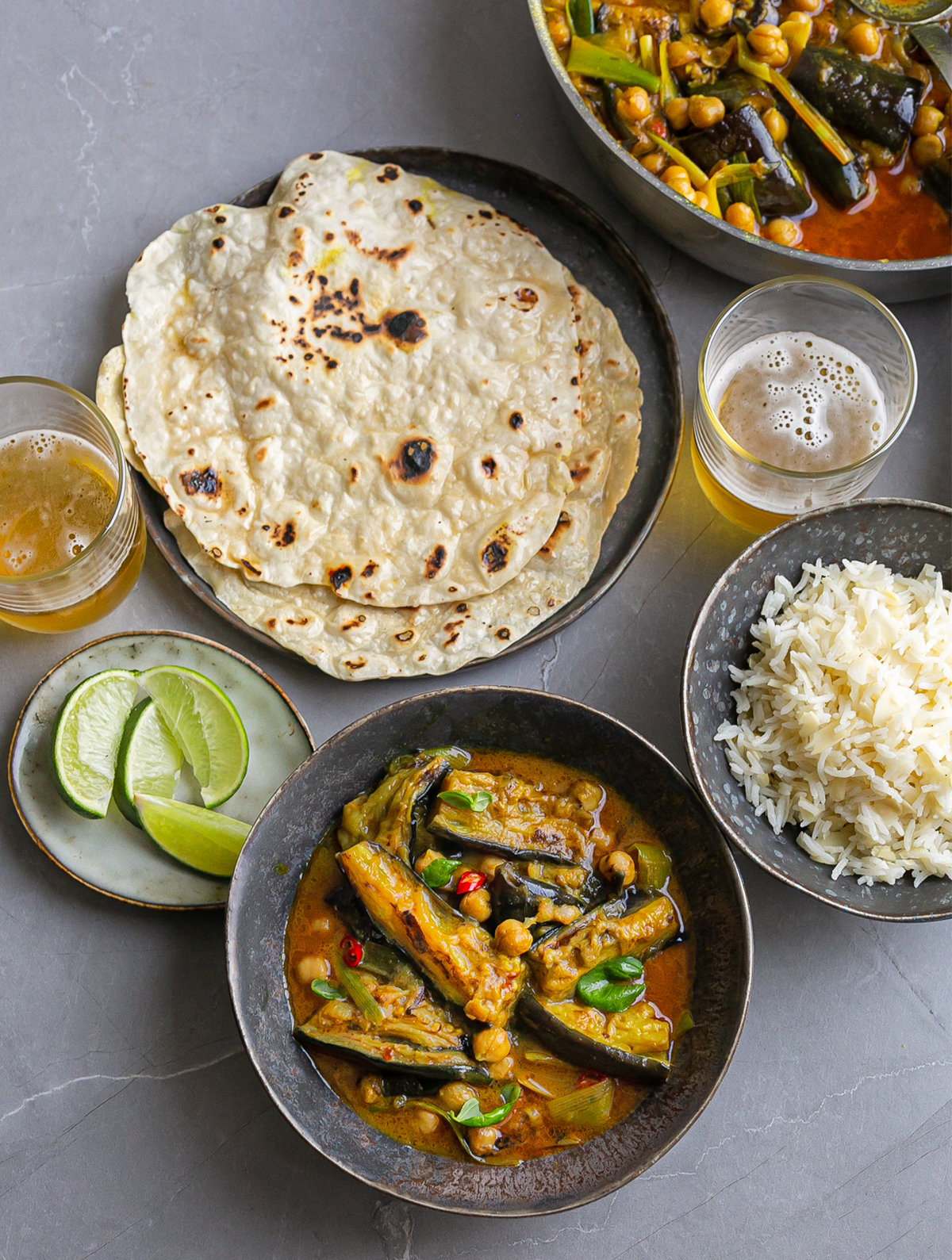 light blue ceramic bowl filled with Fragrant Aubergine Coconut Curry next to rice, flatbreads and a beer