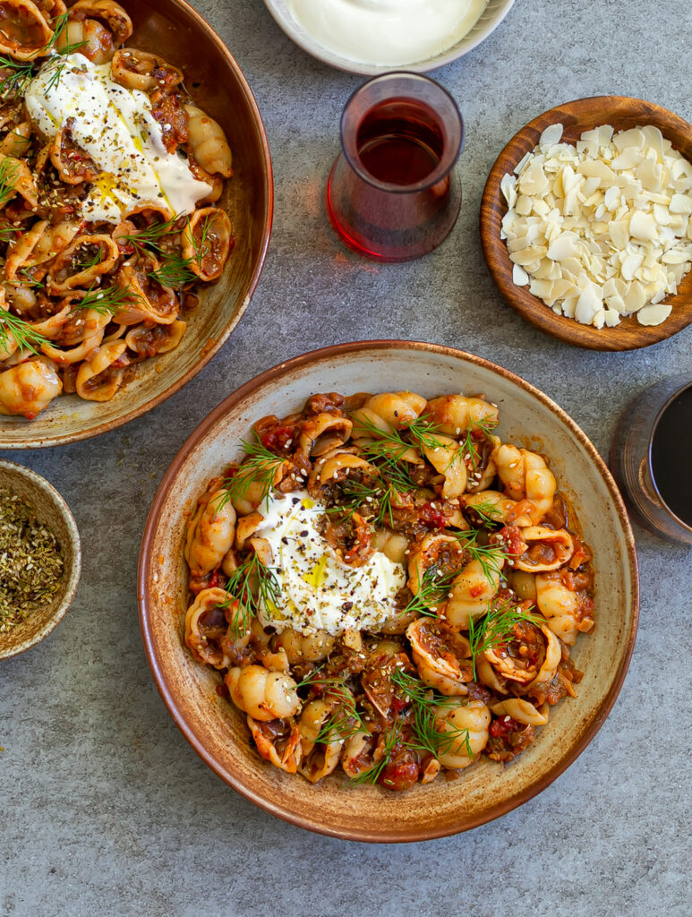 brown ceramic bowls filled with Spicy Aubergine Caviar Pasta, topped with yoghurt, dill and za'atar next to glasses of red wine, olive oil and flaked almonds