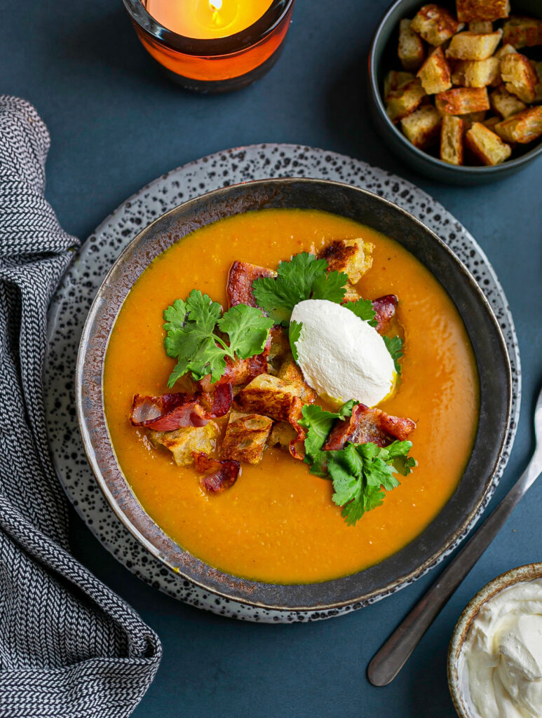 Dark ceramic bowl filled with Hokkaido pumpkin soup and a garnish of crispy bacon, buttered croutons, Greek yoghurt and fresh coriander